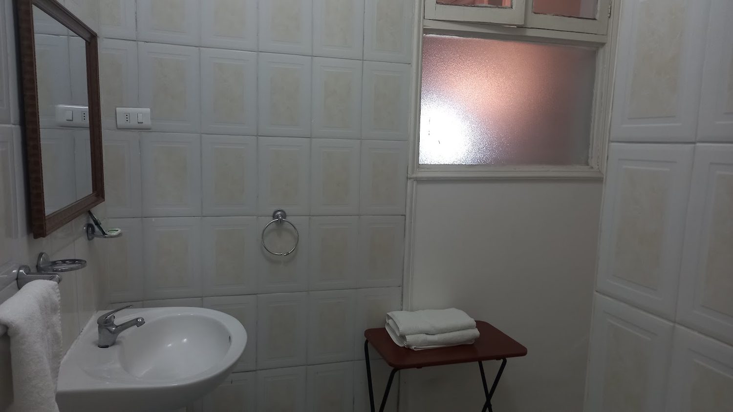 Baño privado del hotel, revestido en cerámica clara, con lavamanos y toallas dobladas